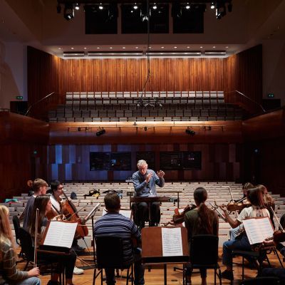 A man with grey hair and a blue jumper conducts an orchestra